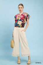 A woman wears oatmeal linen wide leg cropped pants with lace trim paired with a vibrant floral blouse