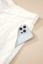 A sleek silver iphone 13 pro with triple lens camera peeking from white fabric near flowy wide leg pants with drawstring waist