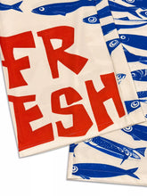 Cream-colored kitchen towel with bold red fresh lettering and blue fish pattern alongside plus size printed wide leg pants