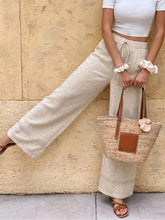 Beige woven straw tote bag with brown leather handles and pink fabric flower paired with casual wide leg pants