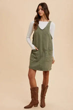 Olive green annie wear adjustable strap denim overall dress with two front pockets worn over a white long sleeve top