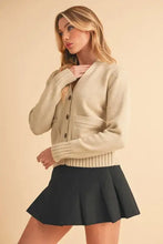 Cream cardigan with ribbed hem button closure, knit pockets, and dark green pleated skirt