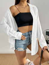 White oversized button up long sleeve shirt worn open over a black crop top and denim shorts for casual wear