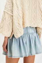 A cream-colored cable-knit sweater with flared sleeves worn over a buttoned ruffled mini denim skirt