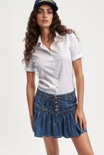 White button-down shirt with short sleeves and rolled cuffs paired with a buttoned ruffled mini denim skirt