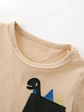Children’s dinosaur pocket t-shirt with beige shirt, black dino peeking from pocket with blue gold spikes. Machine wash cold