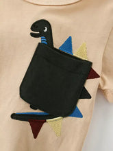 Black embroidered dinosaur with colorful spikes in pocket on beige kids t-shirt, machine wash cold