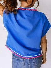 Bright blue short sleeve top with pink pom trim along neckline and hem accented by a white stripe