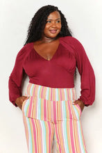 A plus-size woman wearing a maroon satin culture code full size v neck bodysuit with ruched shoulders and striped pants
