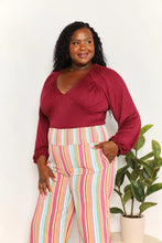 A woman wears a culture code full size v neck maroon satin bodysuit with vibrant wide-leg pastel striped pants