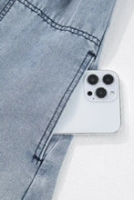 A white iphone 13 pro peeks from light-wash denim jeans pocket worn with a cutout round neck balloon sleeve denim dress