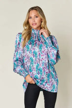 A woman wears a double take full size printed smocked long sleeve blouse with an abstract teal and purple floral pattern