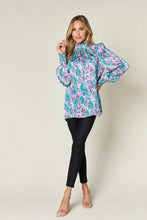 A woman wears a double take full size printed smocked blouse with long puff sleeves in teal purple and white floral pattern