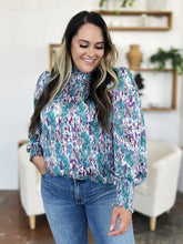 A woman wearing a double take full size printed smocked long sleeve blouse in teal purple and white with puffed sleeves