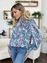 A woman wearing a double take full size printed smocked long sleeve blouse in teal and purple lightweight fabric