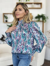 A woman wears a double take full size printed smocked long sleeve blouse with teal and purple abstract pattern