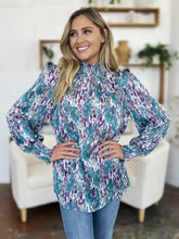 A woman wears a double take full size printed smocked long sleeve blouse with teal and purple abstract patterns on white