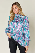 A woman wears a double take full size printed smocked long sleeve blouse with a teal and purple floral pattern