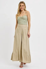 A woman wearing wide leg beige shirring detail pants paired with a sage green lace bodice top with thin straps
