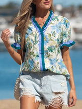 A woman wears a floral print short sleeve blouse with blue green botanical patterns and teal striped trim