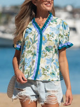 A woman wears a light blue and green floral print short sleeve blouse with ruffled sleeves and striped trim accents