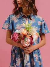A woman wears a blue floral print denim button-down tiered mini dress with pink and white flowers and flutter sleeves