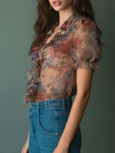 Sheer floral print ruffle trim blouse features ruffled sleeves and neckline in muted pink blue and brown tones