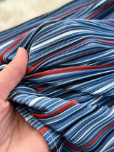 A hand holds a flowing lightweight fabric with bold navy blue white and red diagonal stripes for a striped patchwork denim skirt