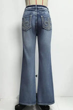 Back view of high-waisted blue denim flare leg jeans with embroidered fleur-de-lis on the back pockets recommend measuring