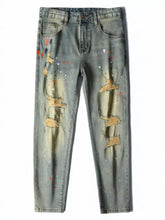 Men’s distressed slim fit jeans in faded blue denim with knee rips reinforced by tan patches and paint splatter accents