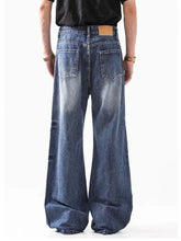 Men’s washed wide leg jeans in faded blue denim with visible stitching and tan leather back pocket patch shown from back side