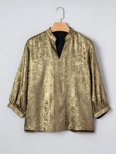 Notched 3/4 sleeve blouse in shimmering gold metallic fabric with subtle texture displayed on wooden hanger