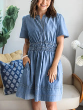A light blue denim dress with notched short sleeves, smocked waistband, and subtle stitching details