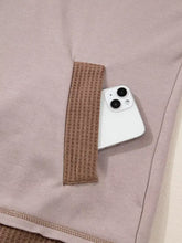 A white smartphone with dual cameras rests in a mauve fabric pocket of an oversize waffle patchwork exposed seam hoodie