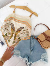 Beige embroidered parrot adjustable strap cami with wooden bead straps and floral patterns on a striped background hanging