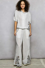 White linen pants with black embroidered floral detailing along the sides and pockets paired with an embroidered floral short sleeve shirt