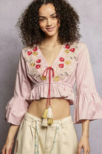 A soft pink cropped blouse with intricate red and gold floral embroidery and flared ruffle sleeves and ruffled hem