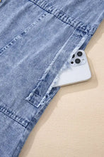 A white iphone with triple camera peeks from the raw hem of a light-wash long sleeve denim dress