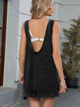Back view of black sleeveless round neck denim overall dress with deep cutout and visible white bra straps
