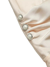 Pearl-encrusted silver buttons adorn the satin fabric of a blush pink ruched spaghetti strap bodysuit