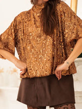 A gold sequined blouse with a high neckline and loose flowing sleeves featuring sequin embellishments and a tie-back design