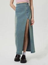 A high-waisted light blue denim skirt with a central front slit and raw hem featuring a zip closure