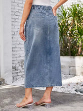 Back view of a mid-blue slit midi denim skirt with waist stretch elastic waistband and two back pockets