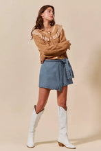 A woman wearing a tan cable-knit sweater with lace detailing and a wrap braided trimmed denim skort tied at the waist