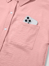 A light pink textured cotton solid color cuffed short sleeve shirt with a front pocket holding a silver iphone
