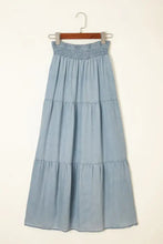 A light blue tiered denim maxi skirt with an elastic smocked waistband and soft flowing fabric