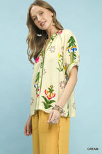 Cream-colored short-sleeved umgee botanical print blouse with vibrant pink blue and green floral embroidery and contrast trim buttons