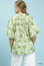 A woman wears an Umgee floral print sage green and white blouse with scalloped edge puff sleeves and high-low hem