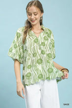 A sage green Umgee blouse with floral print, short flutter sleeves, and scalloped high-low curved hem
