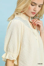 Cream-colored umgee textured henley blouse with waffle-knit fabric, blue embroidery, and ruffle cuffs detail